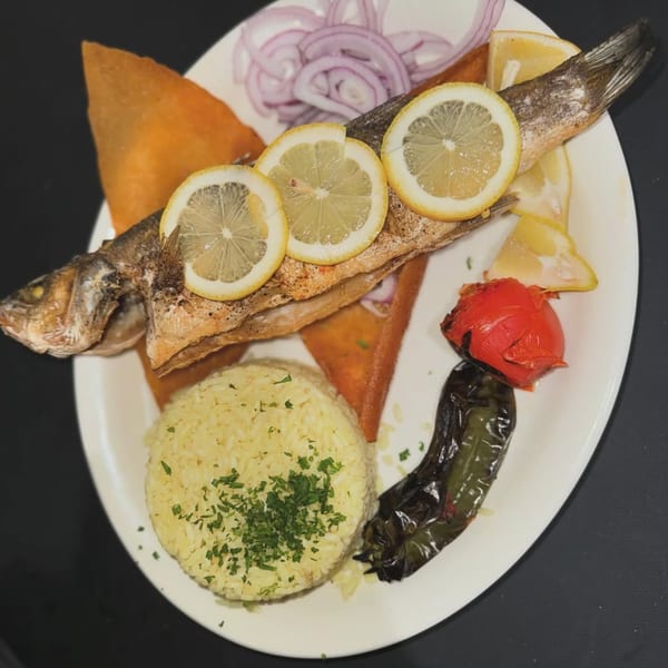 Whole grilled fish with lemon slices, rice, red onions, and roasted vegetables on a white plate.