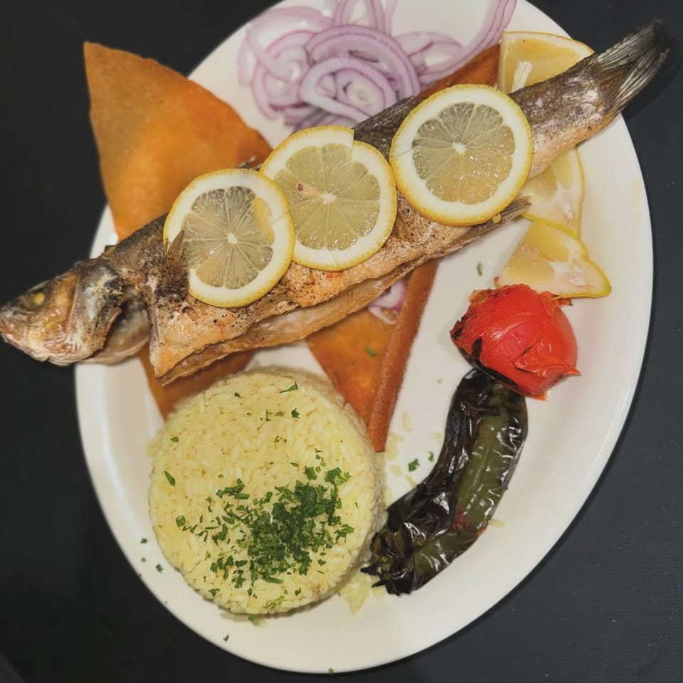 Whole grilled fish with lemon slices, rice, red onions, and roasted vegetables on a white plate.