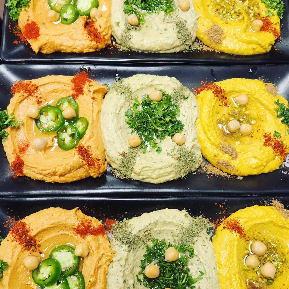 Three rows of hummus platters, each with three distinct flavors and toppings, on black plates.
