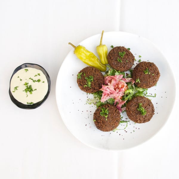 Five falafel patties with pickled peppers, salad, and a side of tahini sauce on a white plate.