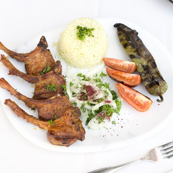 Grilled lamb chops, rice pilaf, charred pepper, tomatoes, and onion salad on a white plate.